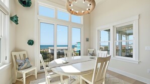 A dining area with a round table, white chairs, a woven ceiling light fixture, and large windows with a view of the ocean. - Ocean Views! One Row to the Beach with Pool & Spa (Oak Island)