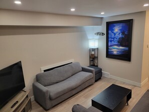Una sala de estar con un sofá gris, una mesa de centro negra, un televisor sobre un mueble de madera, un cuadro colgado en la pared y una lámpara de pie.