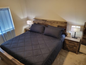 A bedroom with a wooden headboard, a dark quilted bedspread, two bedside tables with lamps, and a window with blinds. - Your SLC Adventure Starts Here • 3BR Townhome • 12 Min to Airport • Sleeps 5+ (West Valley City)