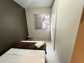 Una habitación de hotel pequeña y estrecha con una cama, una mesita, una ventana con persianas y vistas a edificios.