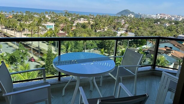 A balcony with a round table and chairs, offering a view of a coastal area with buildings and palm trees.