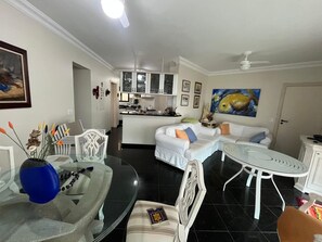 A modern living room with a dining area, white furniture, a kitchen in the background, and a ceiling fan. - Ocean view, 3 bedrooms, beach service and close to everything in Riviera. (Bertioga)