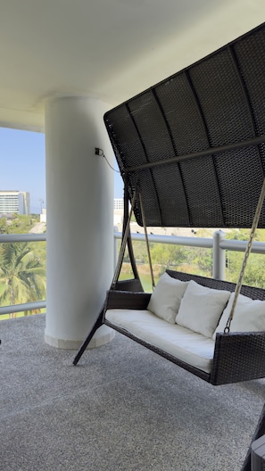 Un balcon agrémenté d’un fauteuil suspendu en osier, d’un pilier blanc et offrant une vue sur des immeubles et de la verdure.