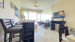 Ein modernes Wohnzimmer mit einer Couch, Sesseln, einem Fernseher und einem Balkon mit großen Fenstern.
