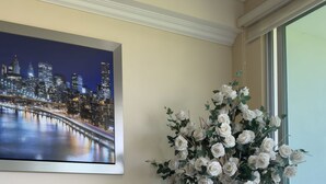 Ein Wohnzimmer mit einem dunkelgrauen Sofa, einer weißen Vase mit Blumen, einem weißen Tisch mit einem kleinen weißen Gegenstand und einem Stadtbild an der Wand.
