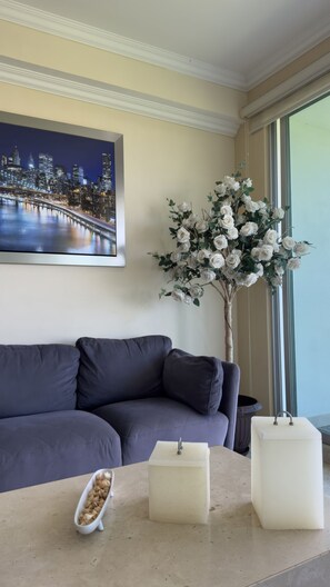 Un salon avec un canapé gris foncé, un vase blanc orné de fleurs, une table blanche sur laquelle se trouve un petit objet blanc, et un tableau représentant un paysage urbain accroché au mur.