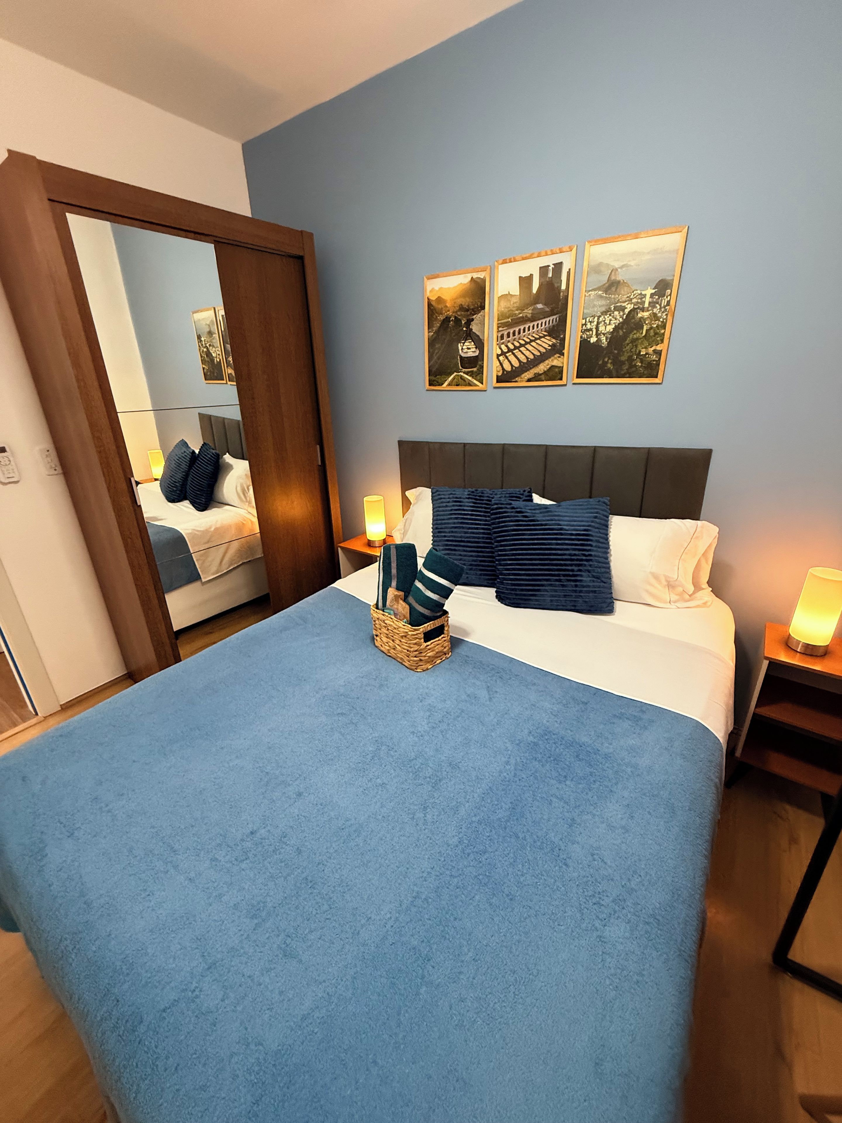 A hotel room with a large bed, a mirror, a nightstand with a lamp, and two wall-mounted pictures.