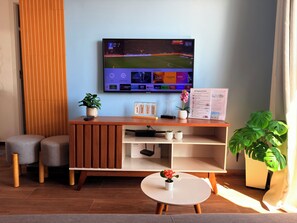 Quarto de hotel moderno com um suporte de TV de madeira, uma TV de tela plana exibindo um canal esportivo, uma pequena mesa com um vaso de planta e uma janela grande com cortinas.