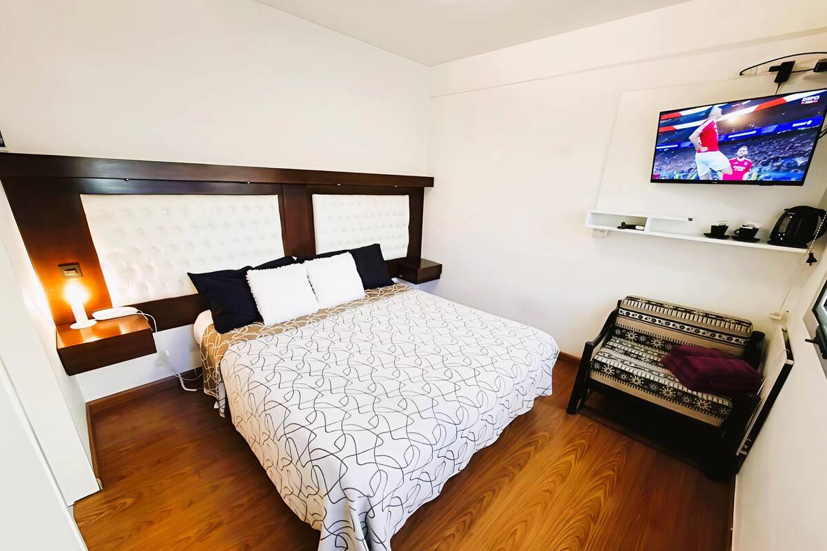 Habitación de hotel con cama, televisor montado en la pared, mesita de noche con lámpara y un estante con objetos.