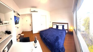 Habitación de hotel con cama, televisión, minibar, ventana con vistas a edificios y puerta.