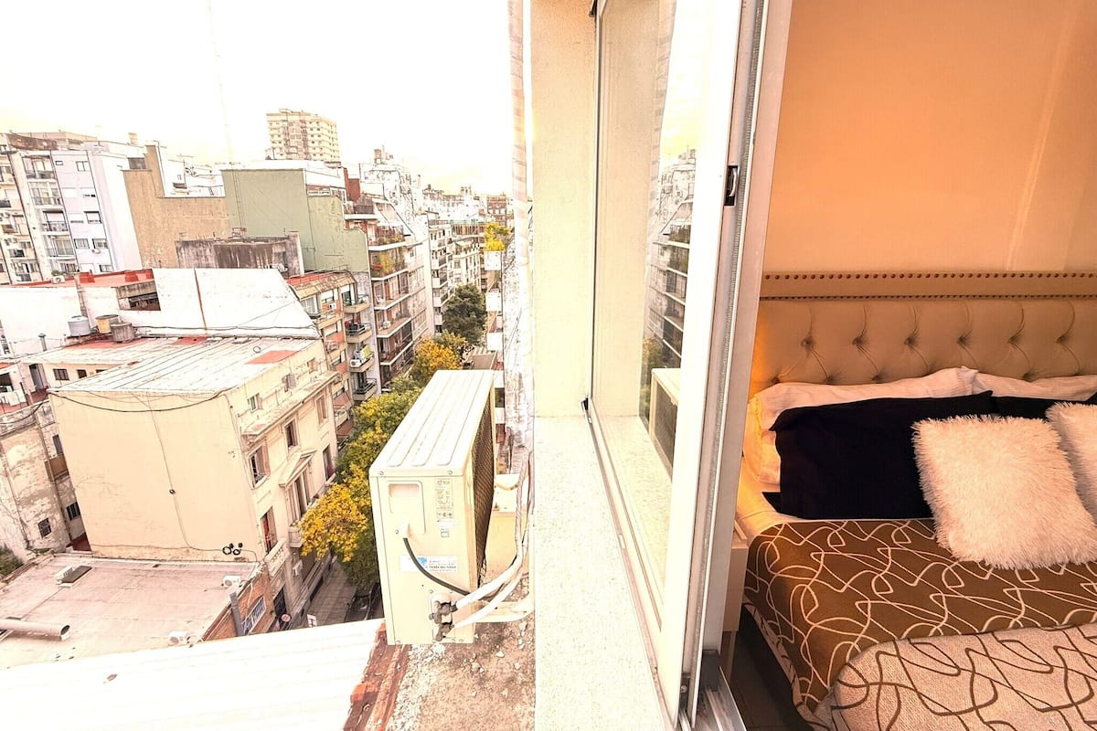 Un dormitorio con cama, ventana y vistas a un paisaje urbano.