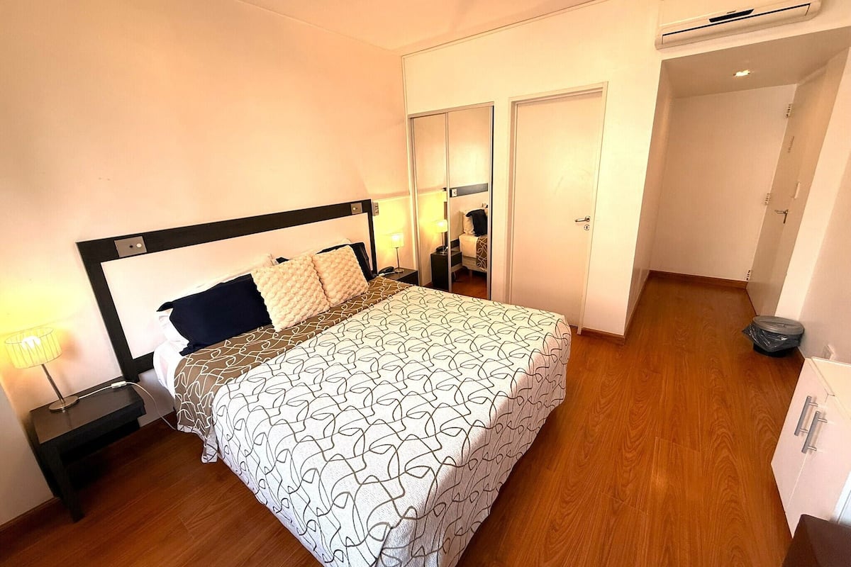 Habitación de hotel con cama, mesita de noche, lámpara, armario y aire acondicionado.