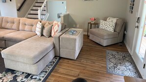 Ein Wohnzimmer mit Ecksofa, Hocker und einem kleinen Beistelltisch mit einer Vase.