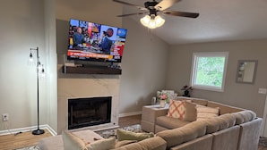 Ein Wohnzimmer mit einem Ecksofa, einem Kamin, einem Fernseher und einem Deckenventilator.