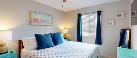 A bedroom with a bed, two bedside tables, a ceiling fan, a window with curtains, and a wall-mounted painting.