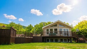 A two-story house with a balcony, a deck, and a view of green trees and mountains.