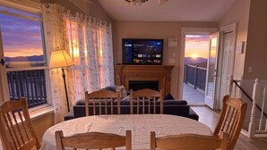 A living room with a fireplace, a television, and a balcony with a view of the sunset.