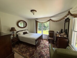Een slaapkamer met een bed, een stoel, een dressoir, een spiegel en een raam met gordijnen.