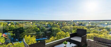A balcony with wicker furniture and a glass table, overlooking a lush green landscape.