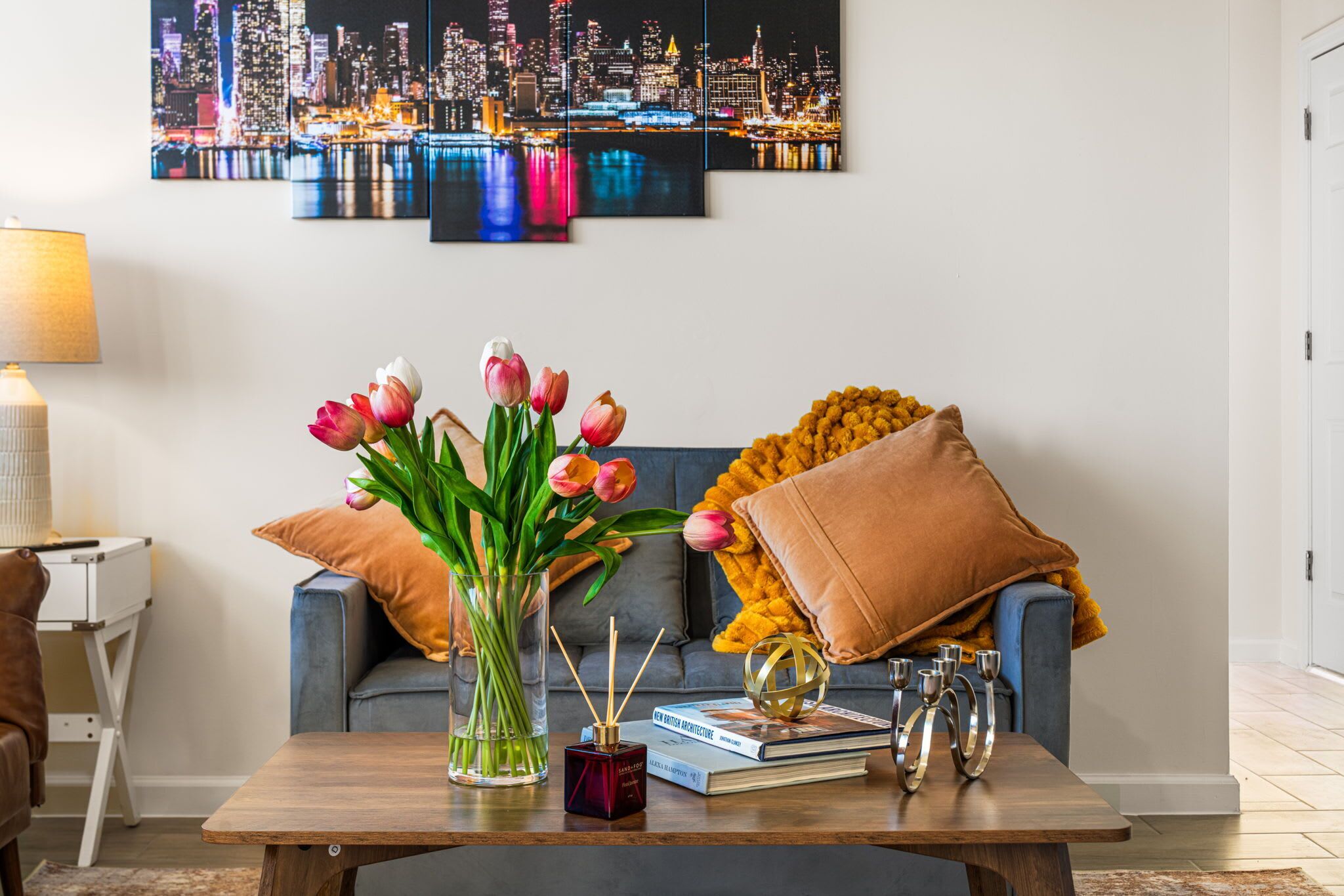 Ein Wohnzimmer mit Stadtbildgemälde, einem blauen Sofa mit brauner Decke, einem Couchtisch mit Büchern und Kerzen sowie einer Tulpenvase.