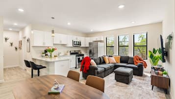 Cocina moderna con armarios blancos, un microondas y un refrigerador. Hay una zona de comedor con una mesa y sillas de madera.