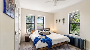 Ein Schlafzimmer mit Bett, Nachttischen, Deckenventilator, Fenstern und Heizkörper.