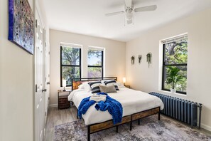 A bedroom with a bed, bedside tables, a ceiling fan, windows, and a radiator. - Bright 2BR w Free Parking, Near NYC (East Orange)