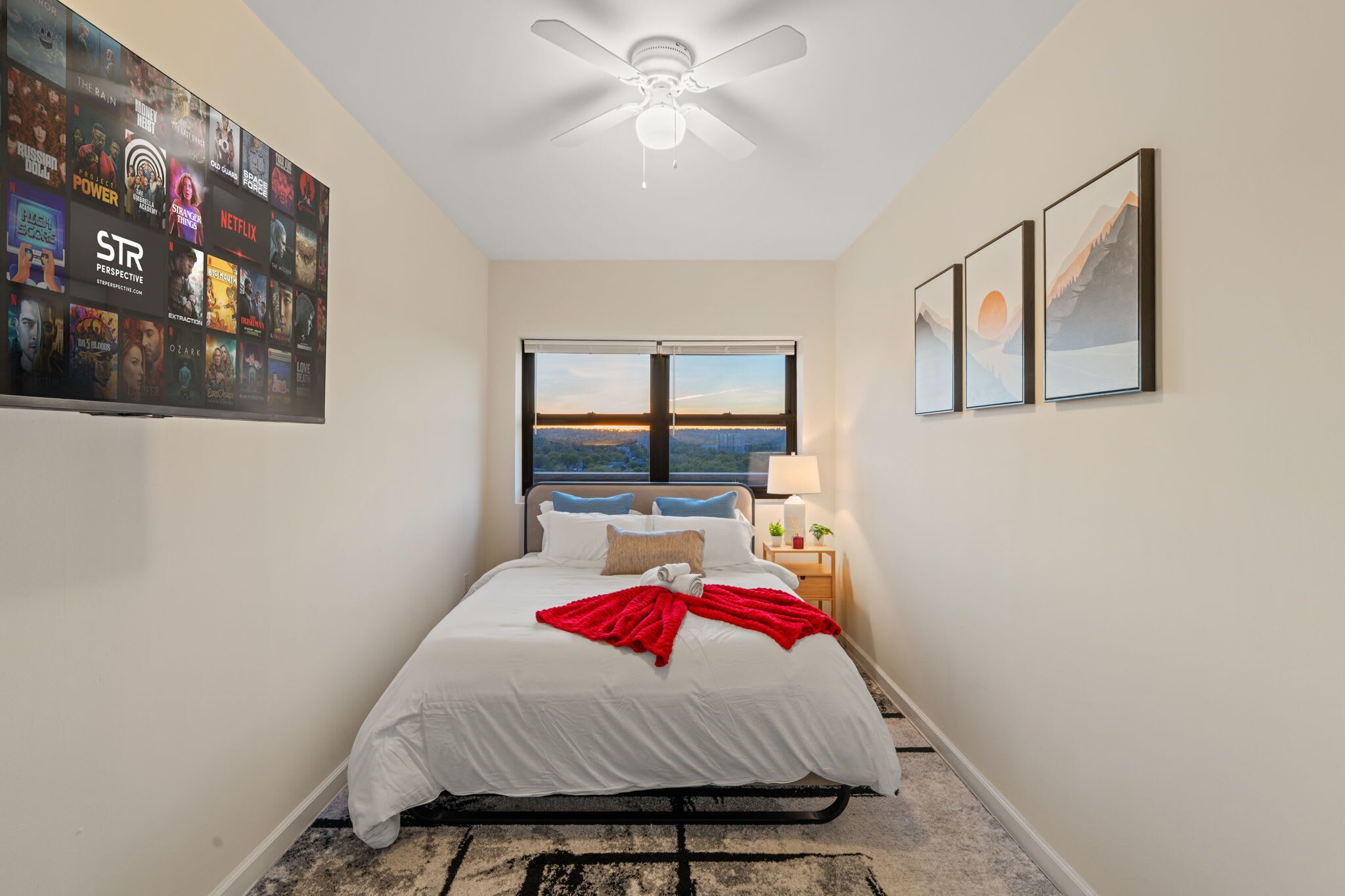 Une chambre à coucher comprenant un lit, un ventilateur de plafond, une fenêtre avec vue et un téléviseur fixé au mur qui diffuse différentes chaînes.