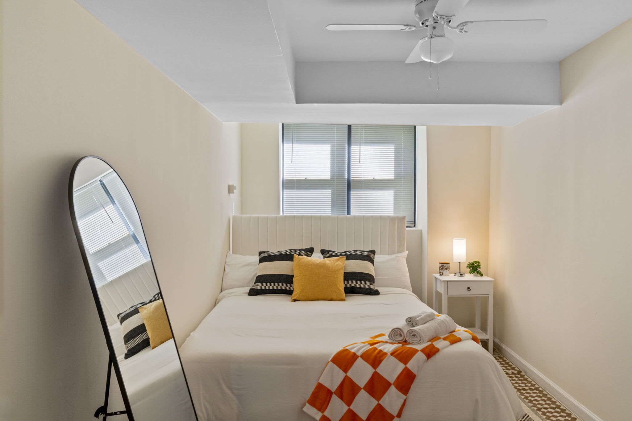 A hotel room with a bed, a ceiling fan, a bedside table with a lamp, and a window with blinds.