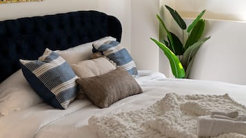 Ein Bett mit dunklem Kopfteil, mehreren Kissen, einer weißen Decke und einer Zimmerpflanze in der Ecke.