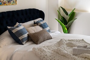 Een bed met een donker hoofdbord, meerdere kussens, een witte deken en een groene plant in de hoek.