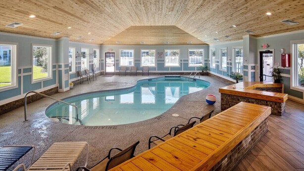 An indoor swimming pool with a wooden ceiling, seating area, and windows. - Flippers Pad at Pebble Beach | Pool View·EI Studio (Emerald Isle)