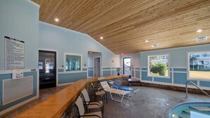 A pool area with a wooden ceiling, a reception desk, lounge chairs, and a pool. - Flippers Pad at Pebble Beach | Pool View·EI Studio (Emerald Isle)