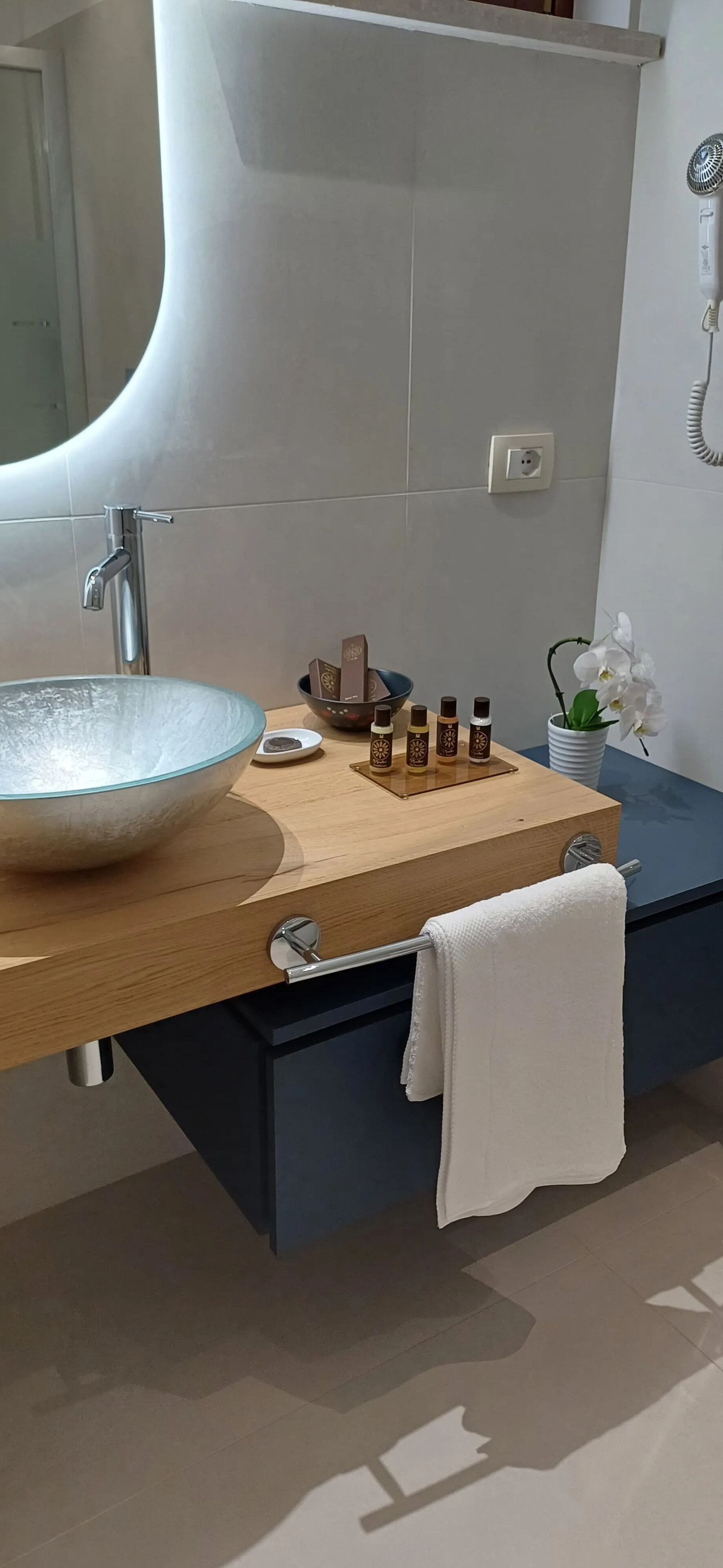 Un baño moderno con un lavabo de vidrio grande, un mueble de tocador de madera, un perchero y una pequeña orquídea en maceta.