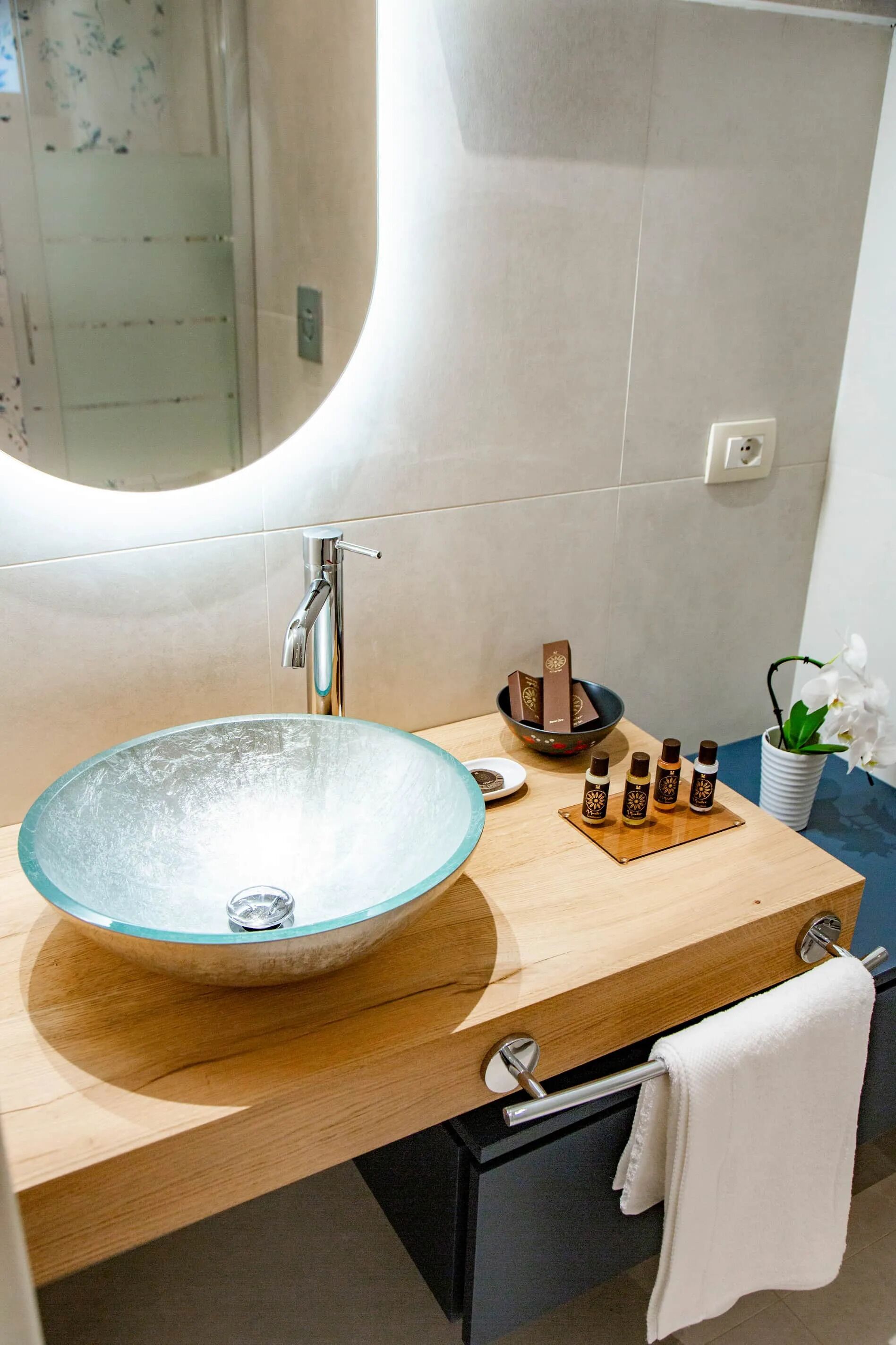 Ein modernes Badezimmer mit einem Waschbecken aus Glas, einem Holzausleger, einem Spiegel, einem Handtuchhalter, einer kleinen Pflanze und einer Ablage mit Dekorationselementen.