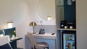 A bedroom with a bed, a desk, a chair, a mirror, a shelf with flowers, and a mini-fridge. - Relais Villa Caia (Noto)