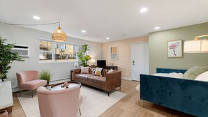 Una sala moderna con un sofá marrón, una cama azul y un sillón rosa. Hay un ventanal grande, una planta y una lámpara colgante.