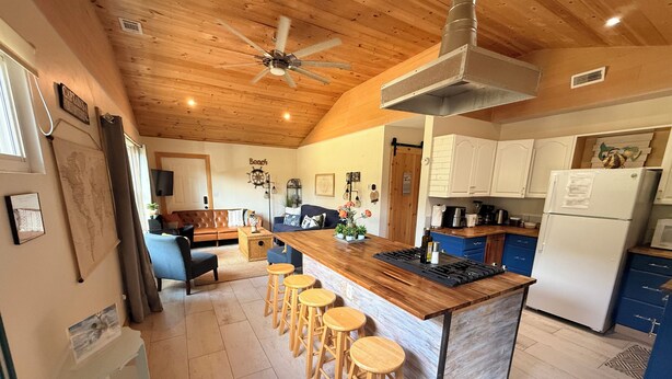 Una cocina con una isla de madera, taburetes de bar, un ventilador de techo y un refrigerador.