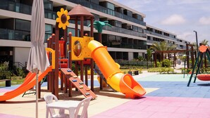 Area giochi per bambini con scivoli e strutture di arrampicata, un tavolo con sedie e un edificio sullo sfondo con diversi balconi.