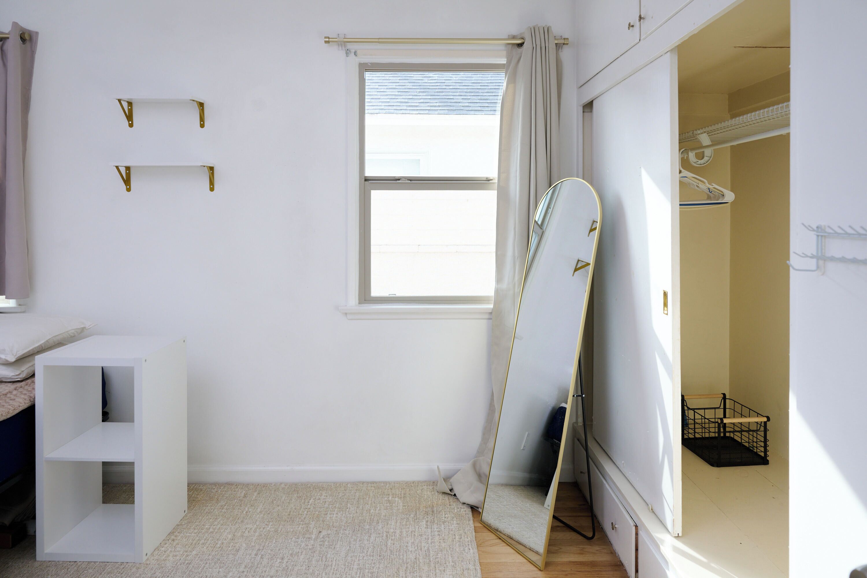 A room with a bed, a white wardrobe, a mirror, a window with curtains, and a small shelf.