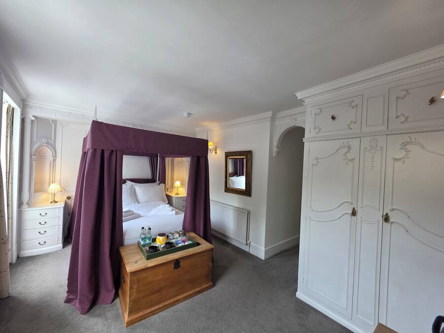 A bedroom with a four-poster bed, a wooden chest of drawers, a wardrobe, and a mirror on the wall.