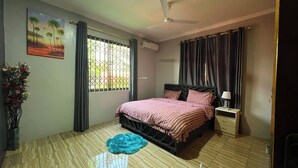 Ein Schlafzimmer mit Bett, Nachttisch, Lampe, Deckenventilator, einem Fenster mit Vorhängen und einer Wand-Klimaanlage.