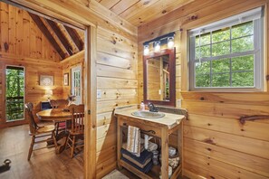A wooden cabin interior with a dining area, a sink with a mirror, and a window with a view of greenery. - Quiet Retreat for Couples, Wi-Fi, Coz,y, Great Location (Rogers)