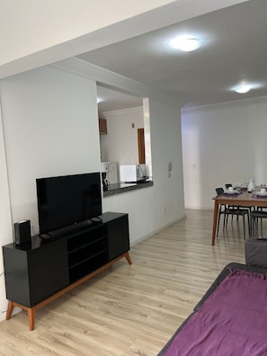 Una sala de estar moderna con un televisor de pantalla plana sobre un mueble negro, una zona de comedor con una mesa y sillas de madera, y una cocina visible a través de un vano abierto.