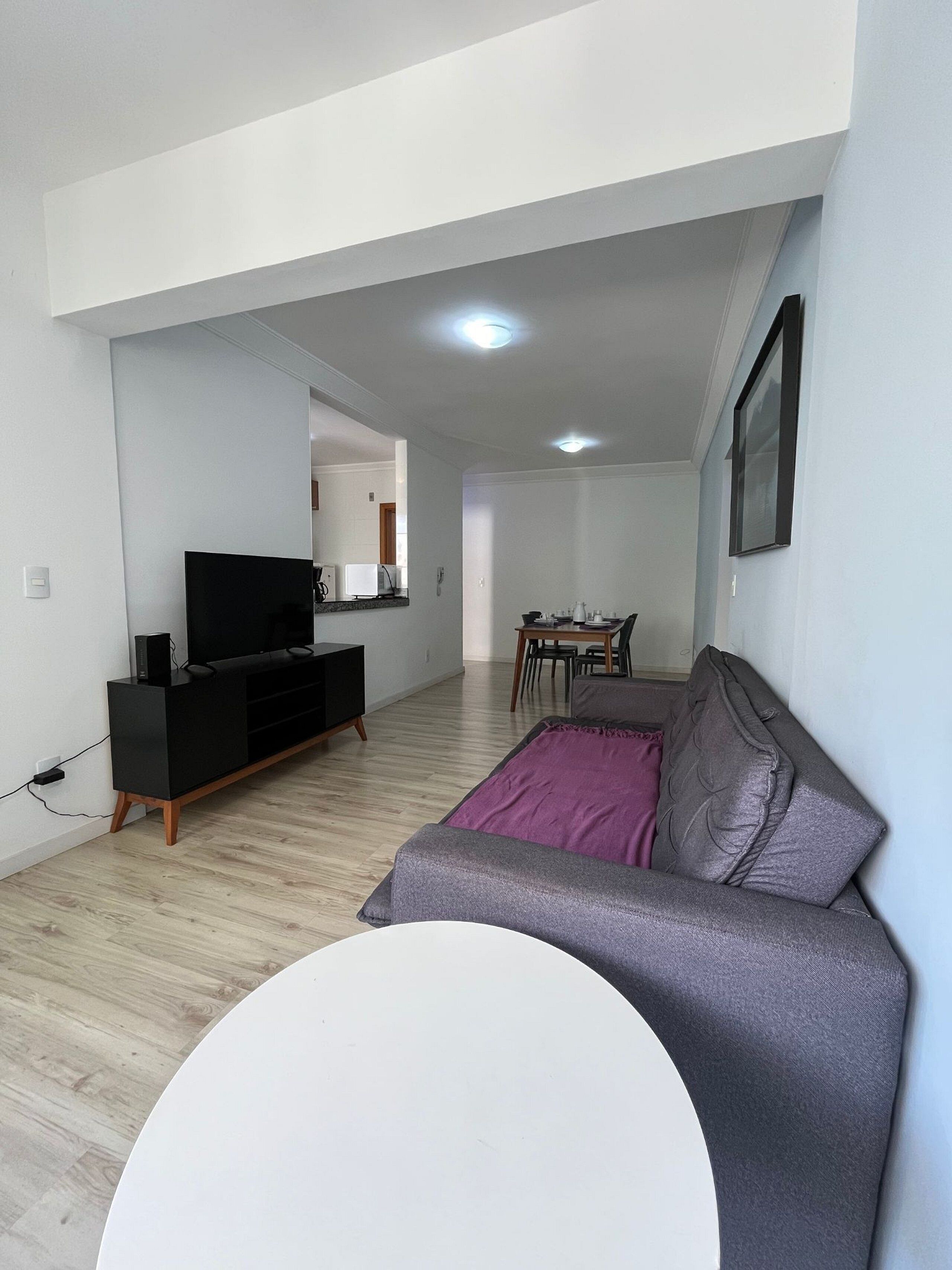 Una sala de estar moderna con un televisor de pantalla plana, un sofá, una mesa de comedor con sillas y una cocina al fondo.