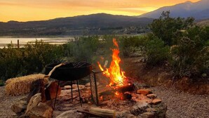 A stone fire pit with a grill, surrounded by a natural landscape with mountains and a body of water in the distance.