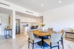 Una cuina moderna amb armaris blancs, una illa central i zona de dinar amb taula rodona de fusta i caires.