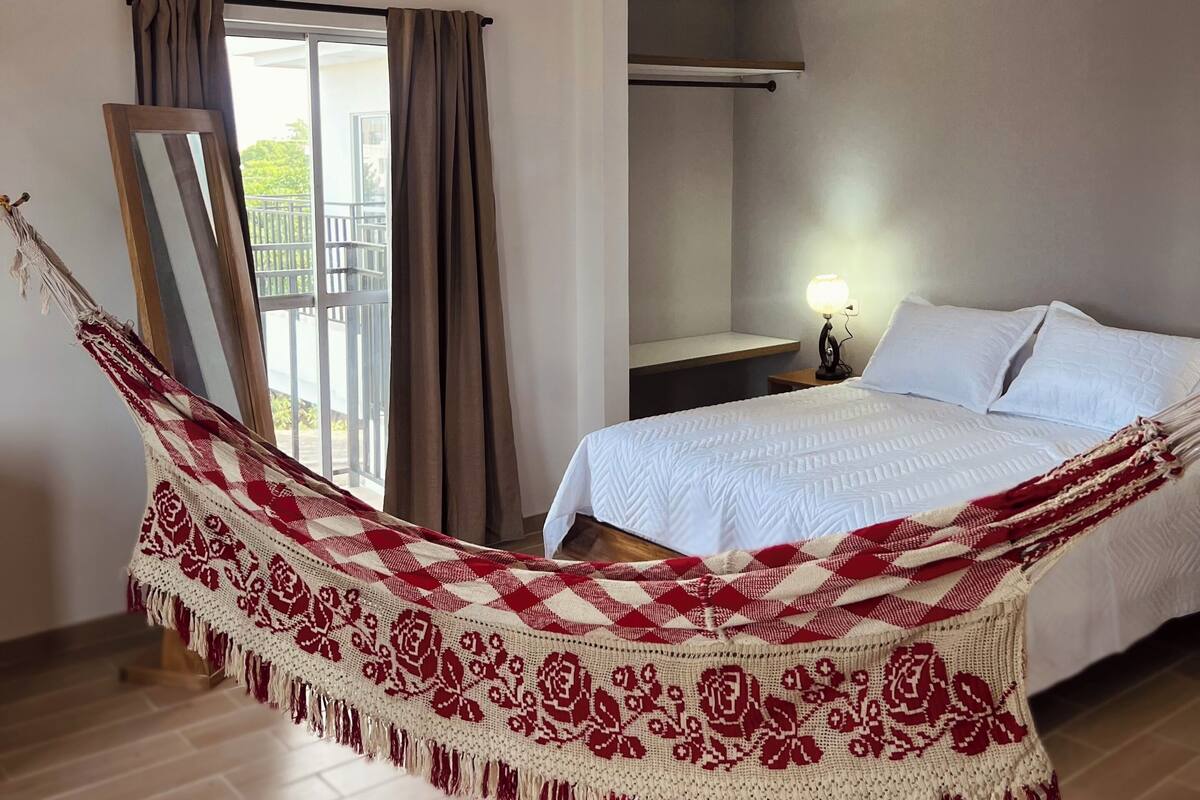 Habitación de hotel con cama, una hamaca, balcón con cortinas y mesita de noche con lámpara.
