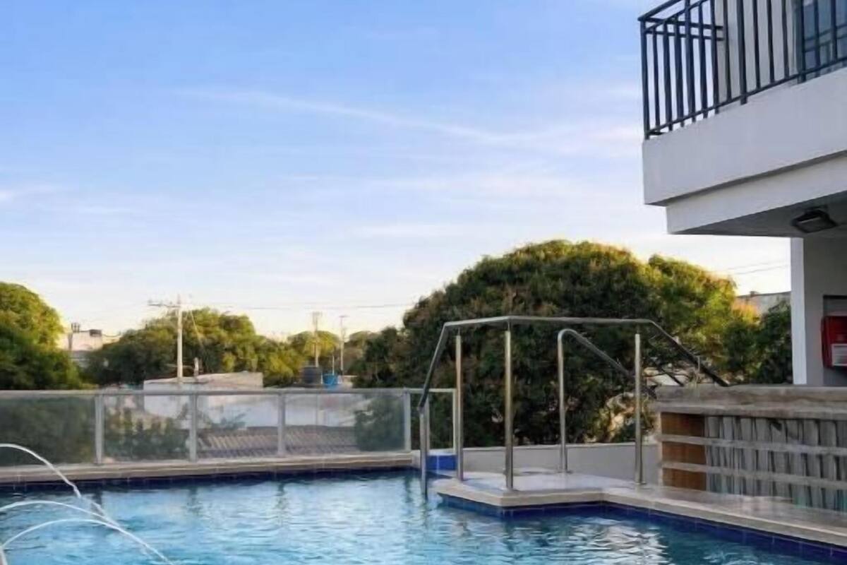 Una piscina con una escalera metálica, rodeada de una valla, y un edificio con un balcón al fondo.