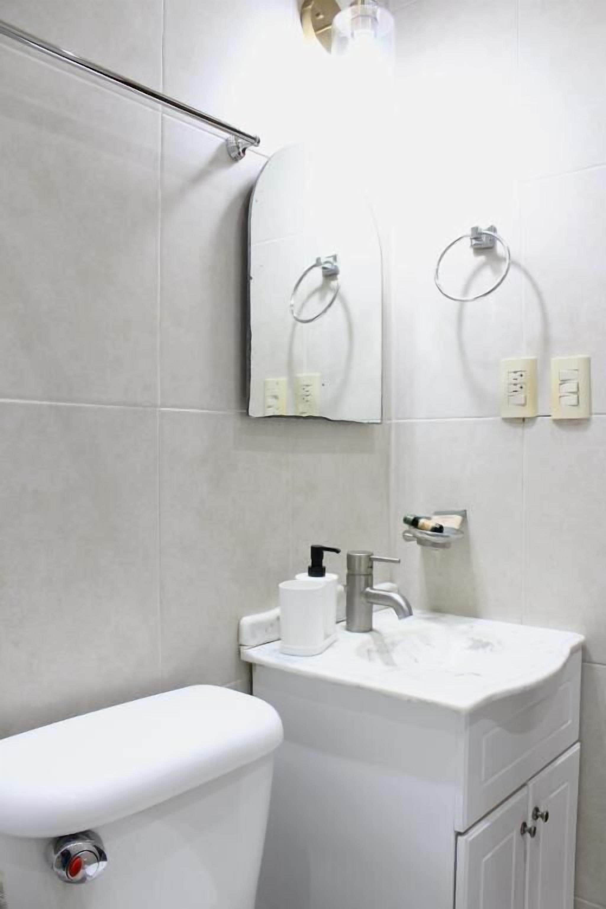 Un baño con inodoro, lavabo con dispensador de jabón y dos anillos para toallas en la pared.